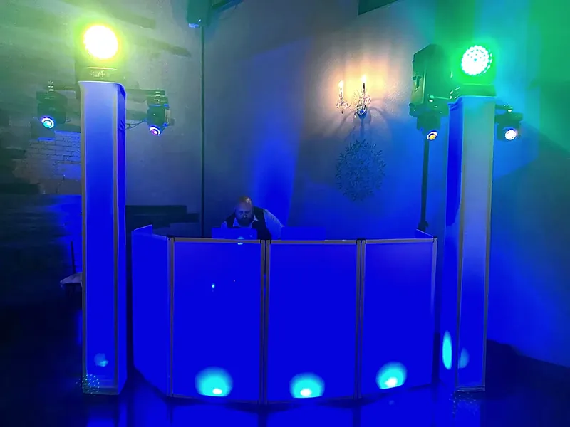 An HPN DJ booth lit blue and flanked by two uplit columns and four moving-head lights, set up in a venue before guests arrive.