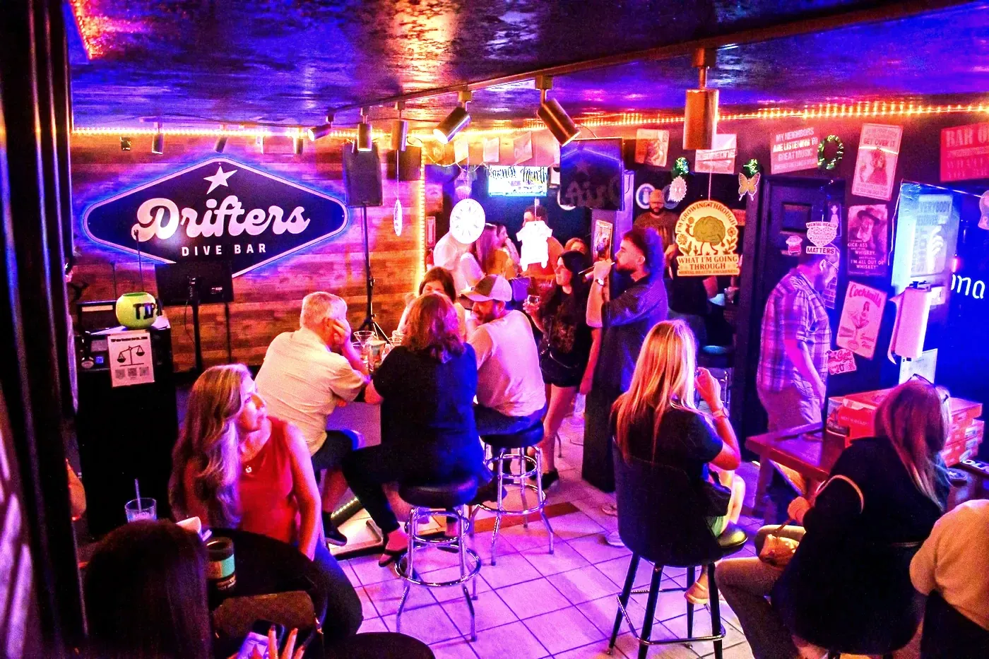 A busy neighbourhood bar with guests at high tables under coloured lighting.