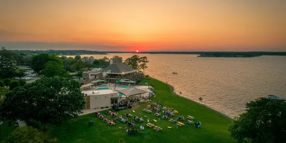 An aerial view of Breakwater Grill's lakeside grounds at sunset, with guests seated at picnic tables beside the water.