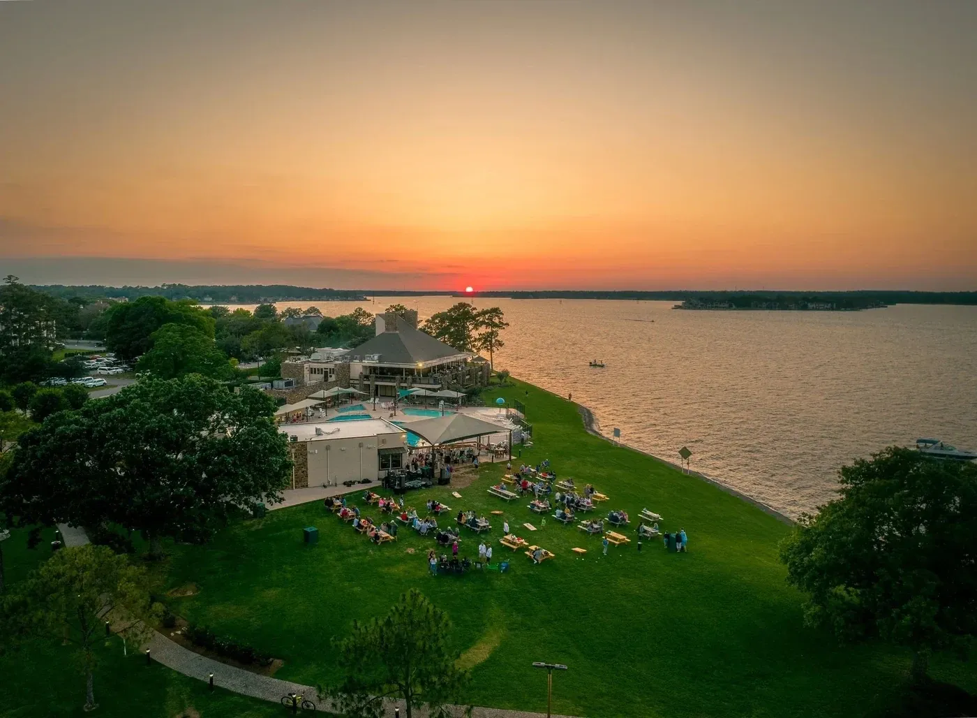 An aerial view of a lakeside venue at sunset, with guests at picnic tables on the lawn beside the water.