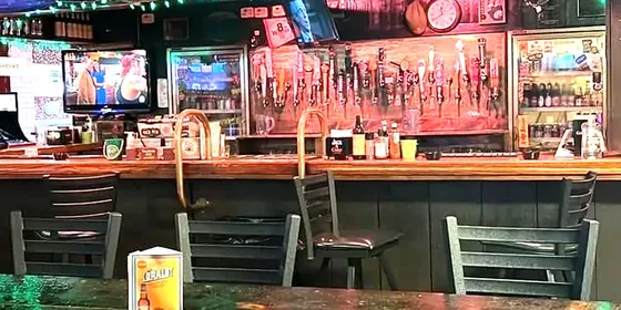The bar of an Irish pub: an empty high table in front of a wooden counter with a long row of beer taps, under red lighting and a string of green bulbs.