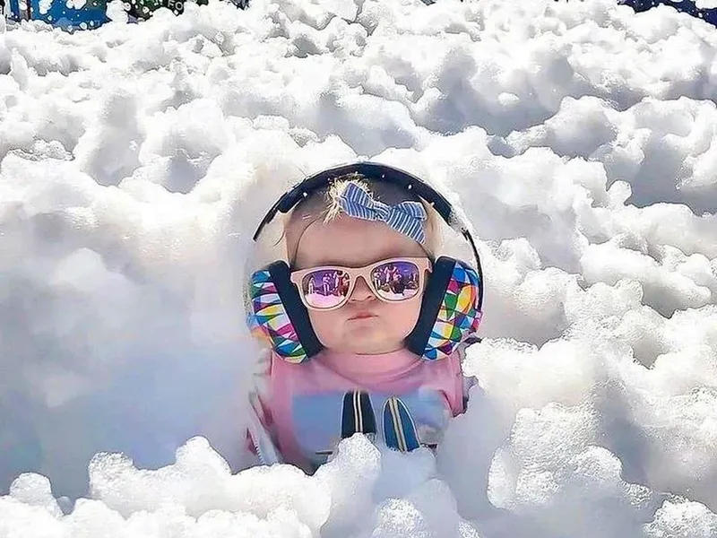 A small child in ear defenders and sunglasses sitting in deep white foam.