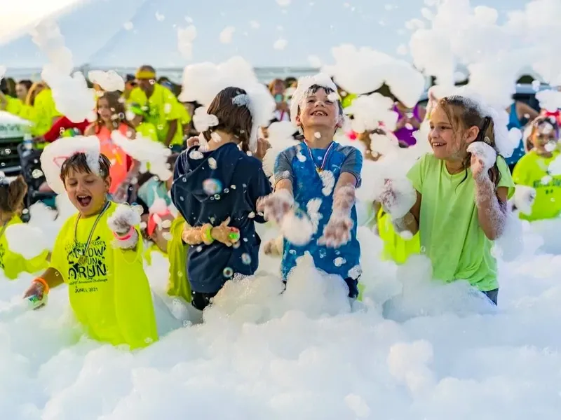 Children in matching event shirts laughing and throwing foam at an outdoor community event, with a marquee and parents behind them.