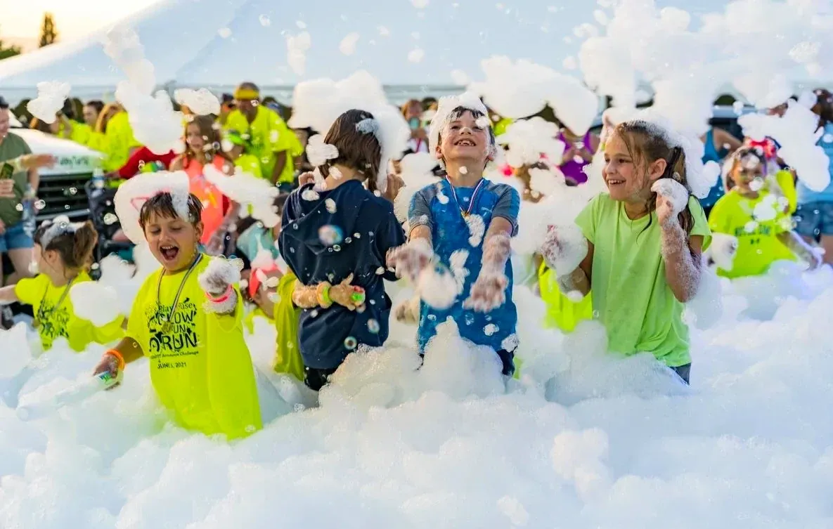 Children in matching event shirts laughing and throwing foam at an outdoor community event, with a marquee and parents behind them.