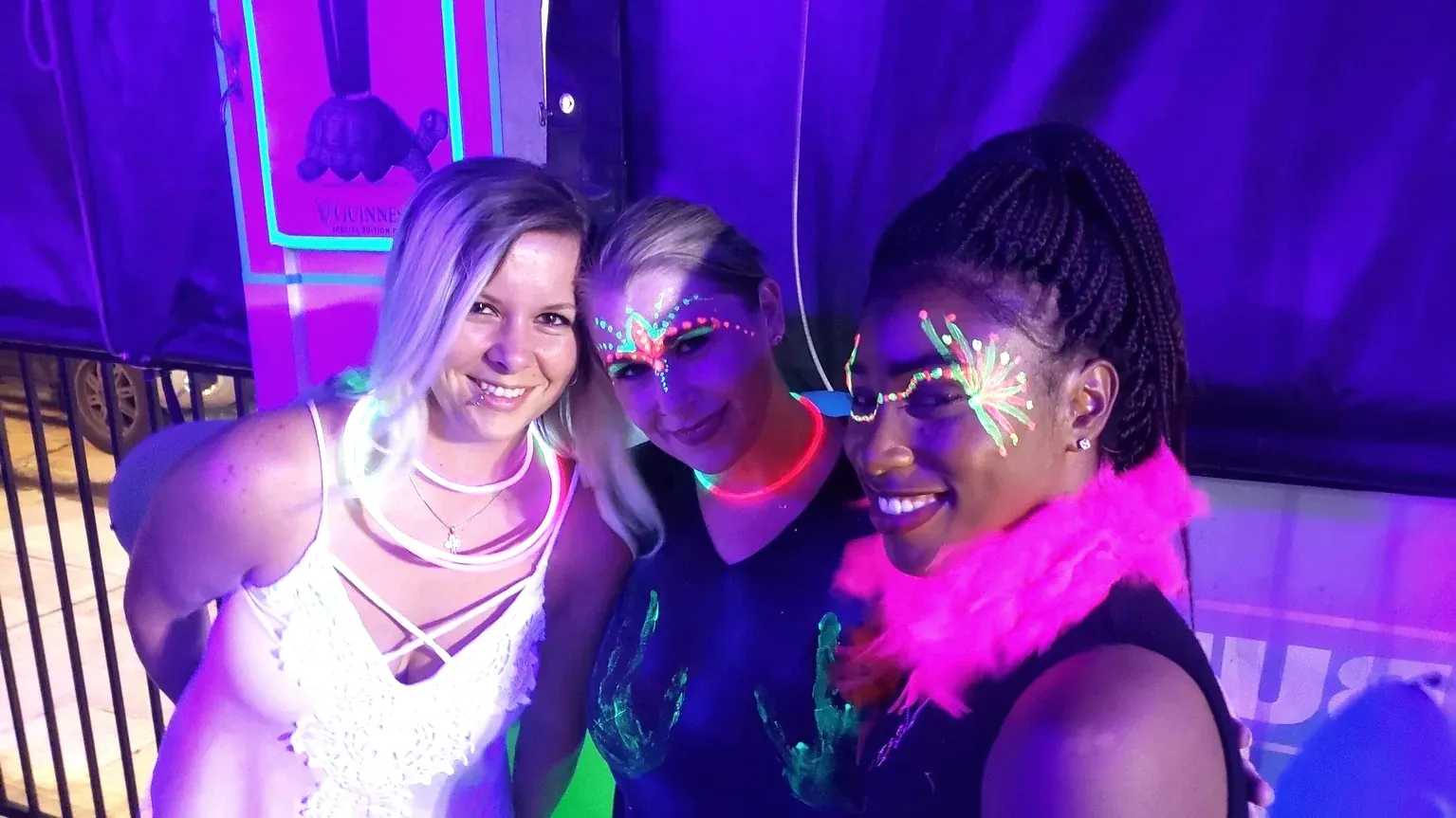 Three guests in glowing face paint and neon accessories at a blacklight party.