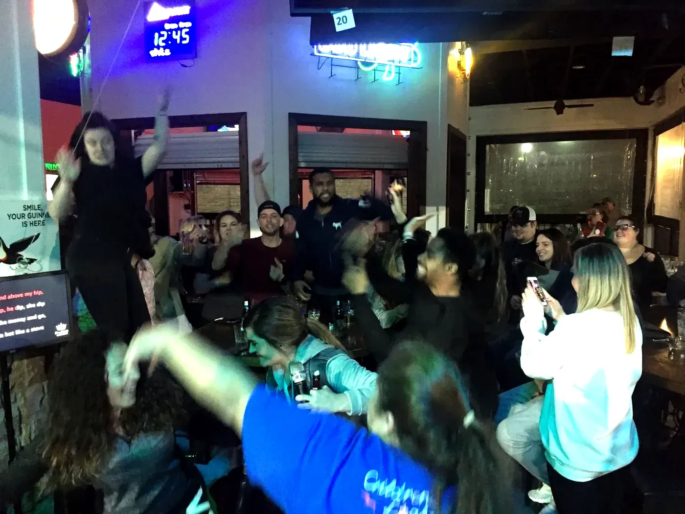 A packed neighbourhood bar with guests on their feet, singing along on a karaoke night.