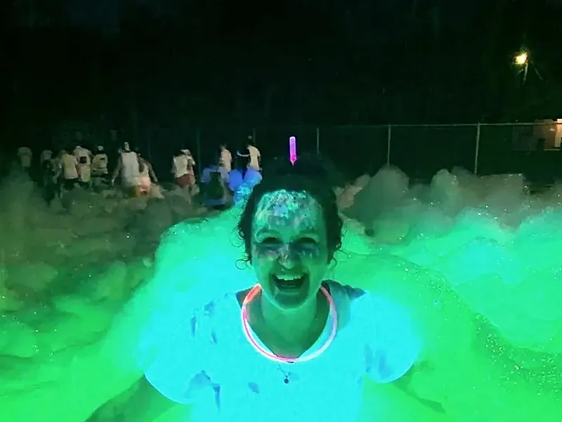 A guest laughing in chest-deep foam at night, lit green by UV lighting, with a crowd behind.