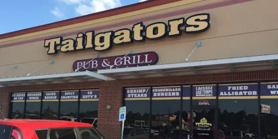The front of Tailgators Pub & Grill under a blue sky: the lit sign over the windows, with boards advertising wings, burgers, steaks and fried alligator.